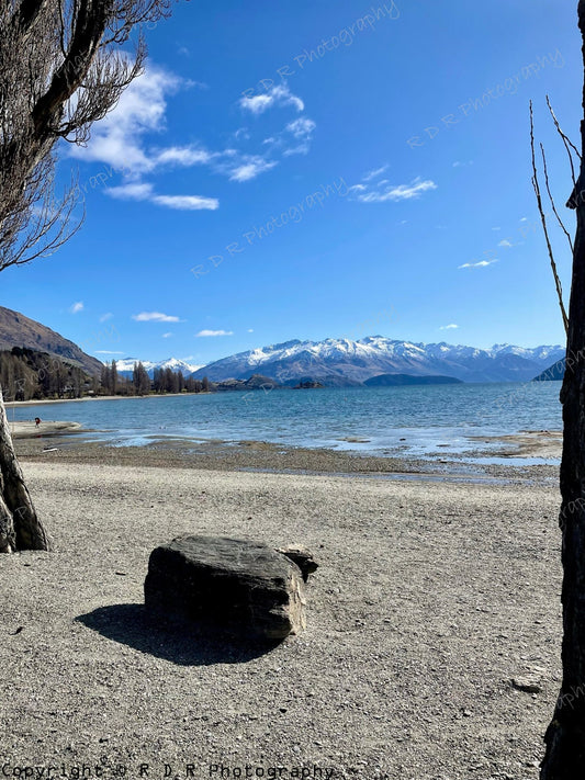 Lake Wanaka shoreline framed by trees, with still water, a solitary rock, and snow-capped mountains beneath a clear blue sky.