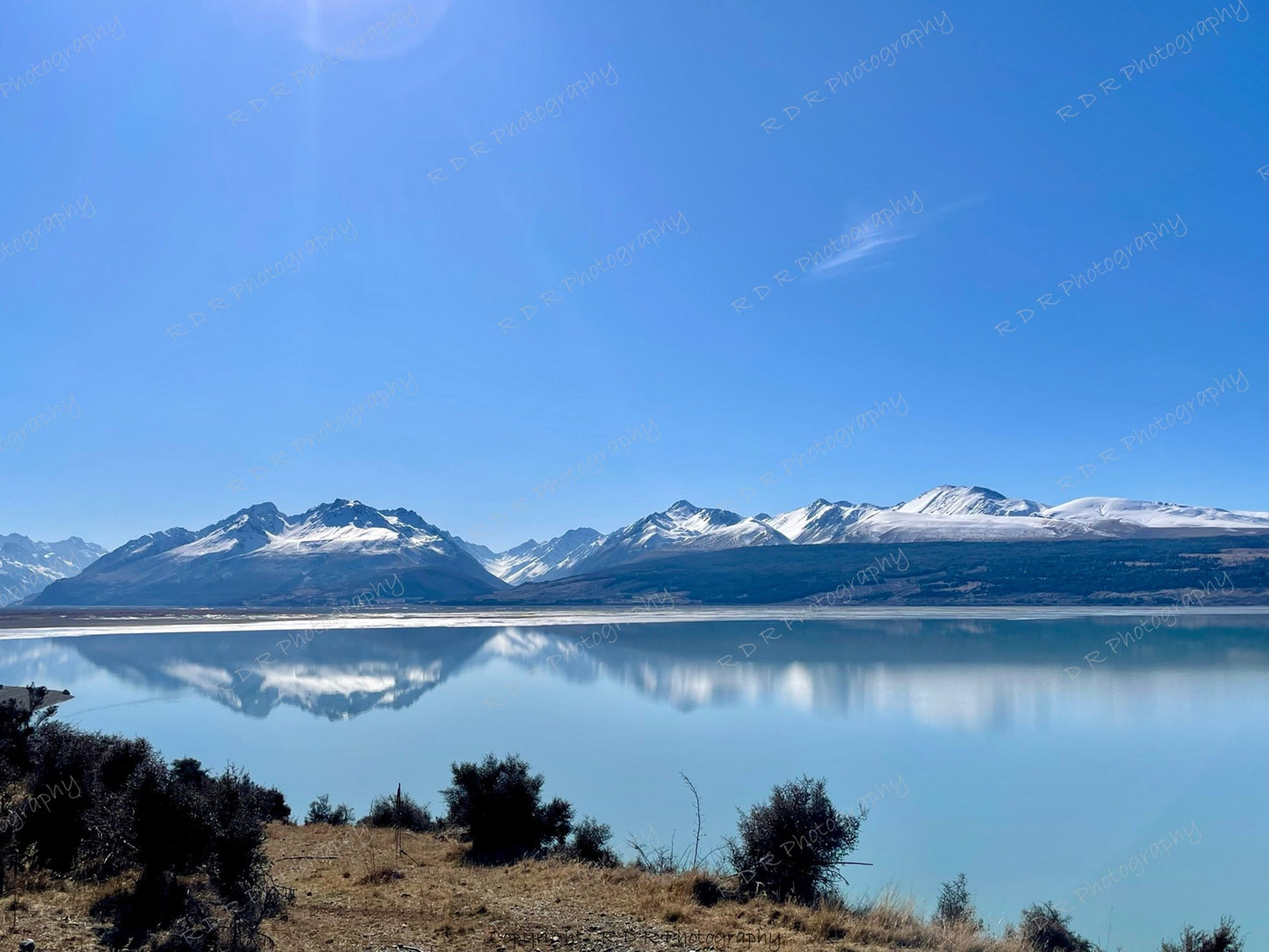 Snow-capped Southern Alps reflected in calm water near Mount Cook under a clear sky, creating a tranquil winter landscape.