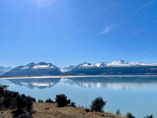 Snow-capped Southern Alps reflected in calm water near Mount Cook under a clear sky, creating a tranquil winter landscape.